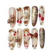 Gingerbread Wishes long almond press-on nail set featuring creamy ivory, champagne gold glitter, gingerbread-inspired nail art, 3D ribbon bows, red candy accents and sparkling stars.